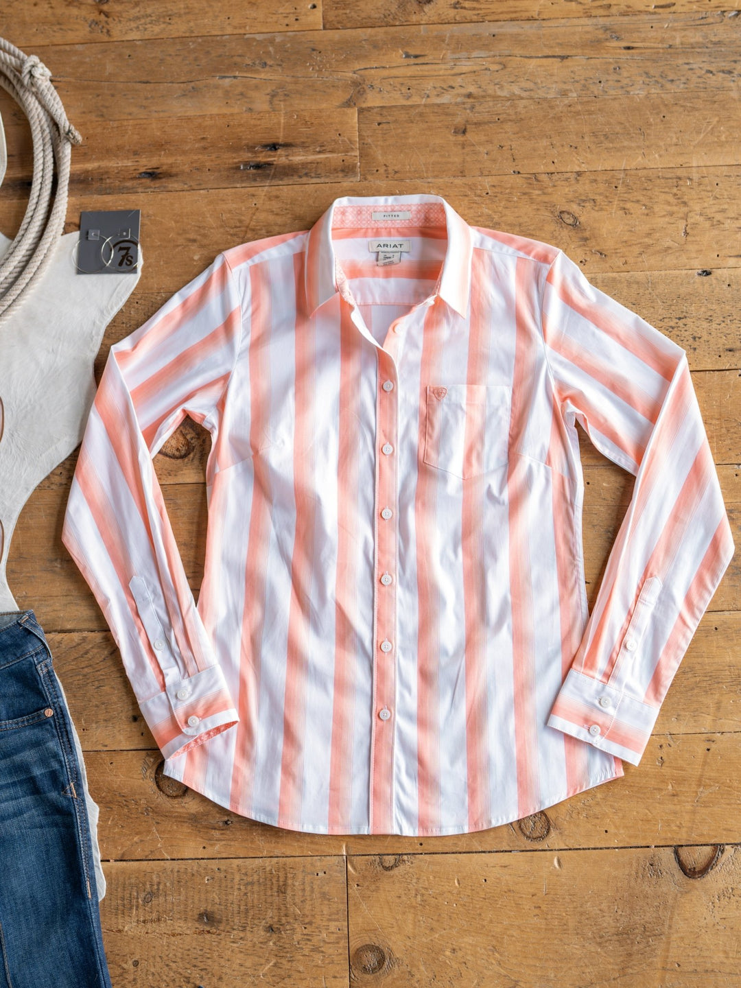 The Forada by Savannah Sevens western life{&}style, a vertical pink and white striped button-up top, is displayed on a wooden floor beside blue jeans and a coiled rope.