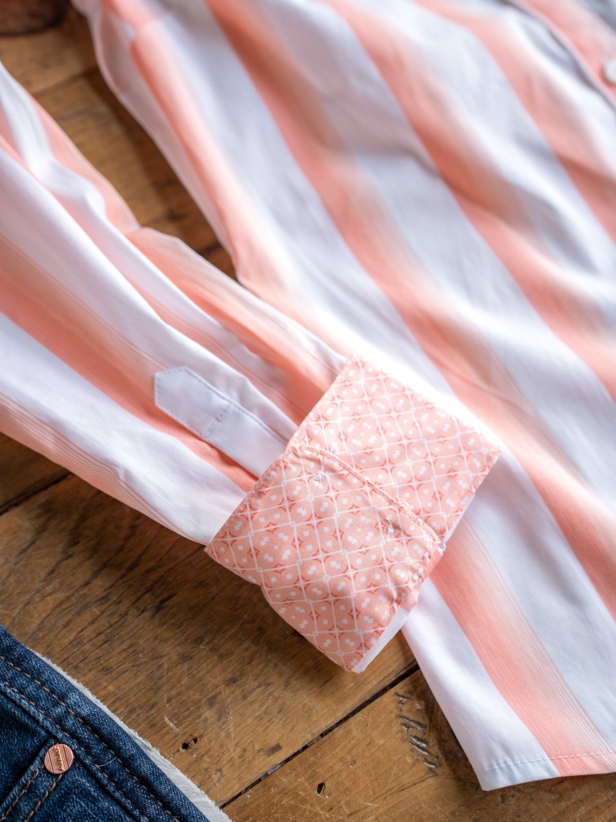 A close-up of The Forada by Savannah Sevens western life{&}style, a stretch button-up with pink and white stripes. Its cuff folds to reveal a pink patterned lining, displayed on wood beside denim fabric.