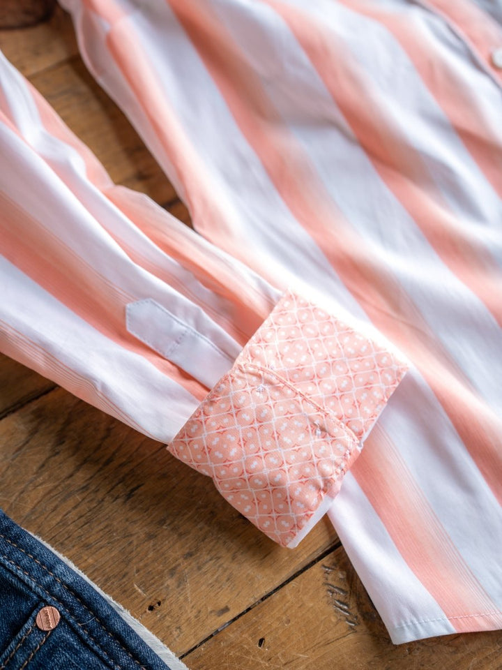 A close-up of The Forada by Savannah Sevens western life{&}style, a stretch button-up with pink and white stripes. Its cuff folds to reveal a pink patterned lining, displayed on wood beside denim fabric.