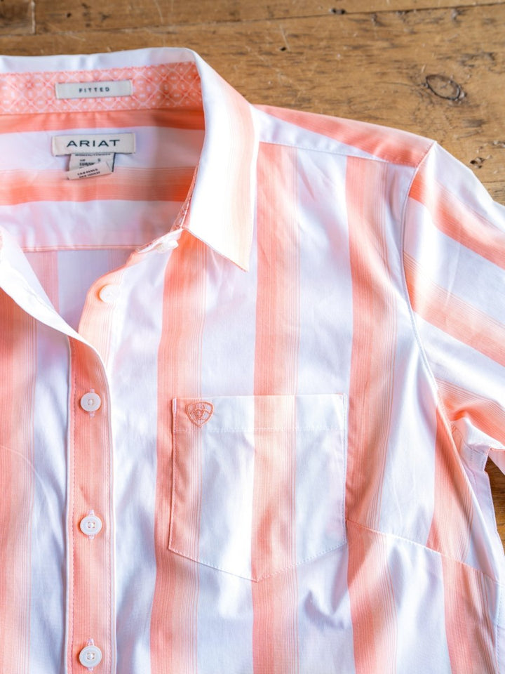 The Forada by Savannah Sevens western life{&}style is a striped button-up top with white and light orange vertical stripes, stretch fabric, collar, front pocket, and inner labels. Laid flat on a wooden surface.