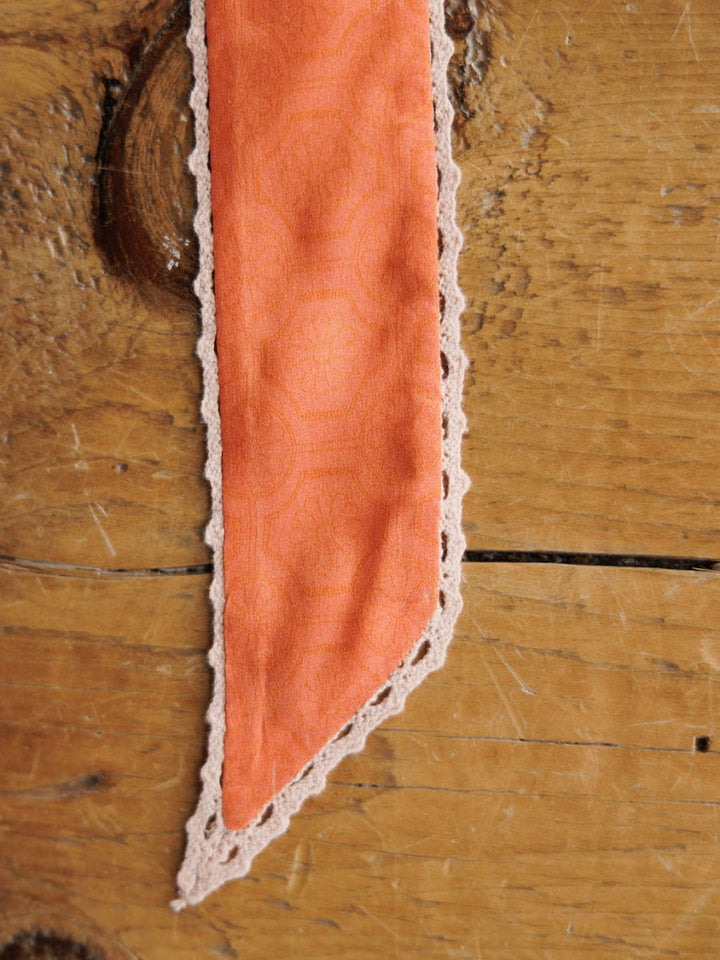 A close-up of the Frankie Twilly by Savannah Sevens western life{&}style, featuring pointed orange fabric with a subtle pattern and crochet trim, displayed on a rustic wooden surface.