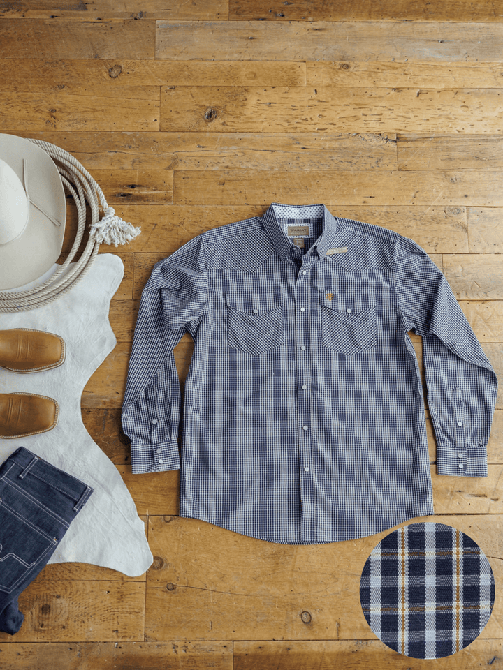 The Freeman Ariat Men's Navy Plaid Pearl Snap shirt by Savannah Sevens western life{&}style is laid flat on a wooden floor with a white hat, brown boots, blue jeans, and rope. A circular inset highlights the plaid snap button detail.