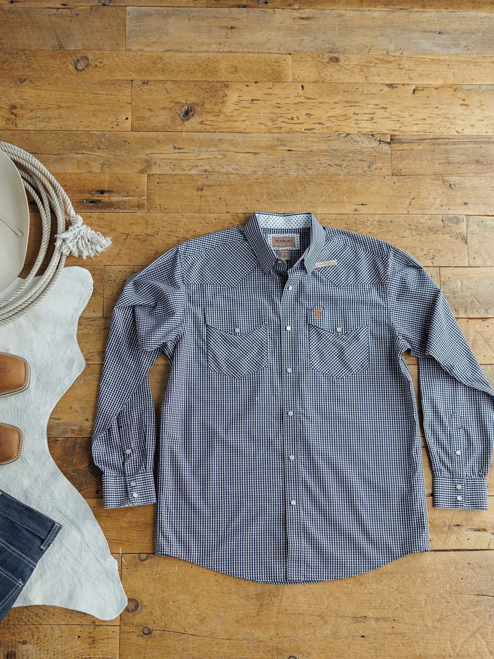 The Freeman Ariat Men's Navy Plaid Pearl Snap shirt by Savannah Sevens western life{&}style is displayed on a wooden floor beside a white cowhide rug and jeans, highlighting classic men's western shirt style.