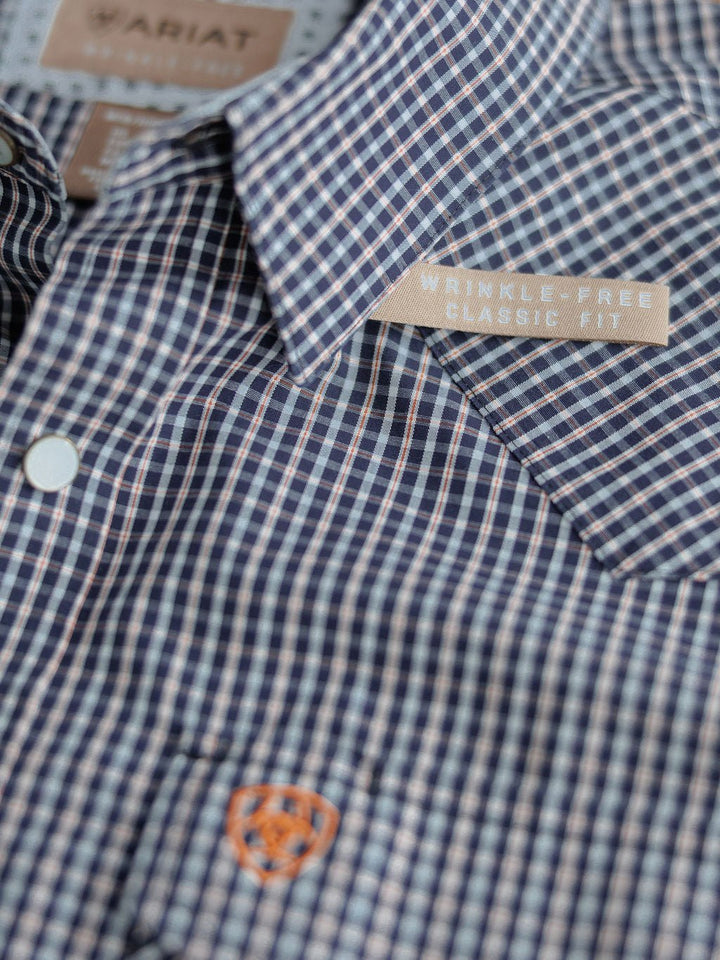 Close-up of the Freeman Ariat Men's Navy Plaid Pearl Snap shirt by Savannah Sevens western life{&}style, featuring a blue and white plaid pattern, pearl snap buttons, and an orange Ariat logo embroidered on wrinkle-free cotton.