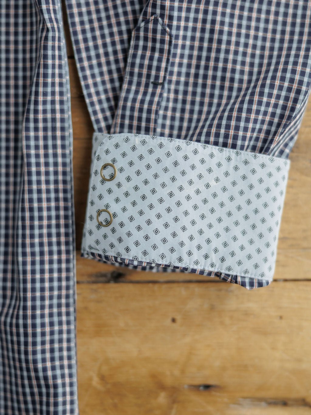 A close-up of the Freeman Ariat Men's Navy Plaid Pearl Snap shirt by Savannah Sevens western life{&}style, showing its blue and white plaid sleeve with a contrasting white cuff featuring a geometric diamond design and two cufflink holes on wood.