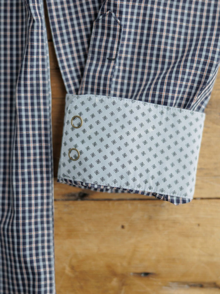 A close-up of the Freeman Ariat Men's Navy Plaid Pearl Snap shirt by Savannah Sevens western life{&}style, showing its blue and white plaid sleeve with a contrasting white cuff featuring a geometric diamond design and two cufflink holes on wood.