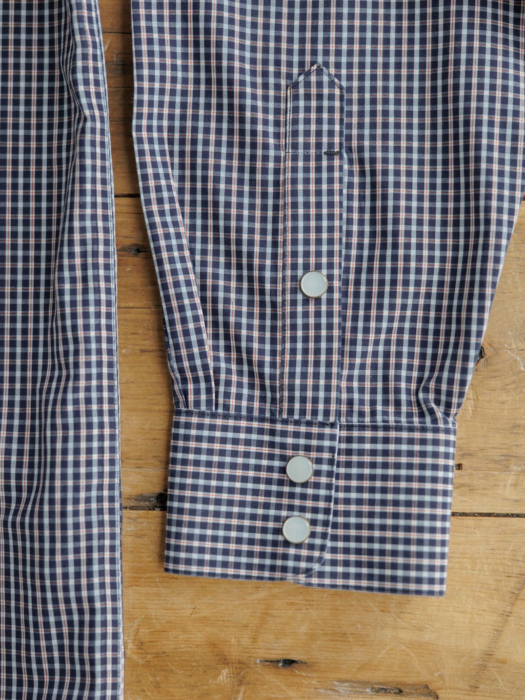 Close-up of a Freeman Ariat Men's Navy Plaid Pearl Snap shirt sleeve by Savannah Sevens western life{&}style, showing three white pearl snap buttons on the cuff, neatly pressed and partially folded on a wooden surface.