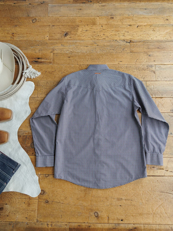 The Freeman Ariat Men's Navy Plaid Pearl Snap shirt by Savannah Sevens western life{&}style is laid flat, back side up, on a wooden floor beside a cowboy hat, cowhide rug, denim jeans, and tan leather shoes.