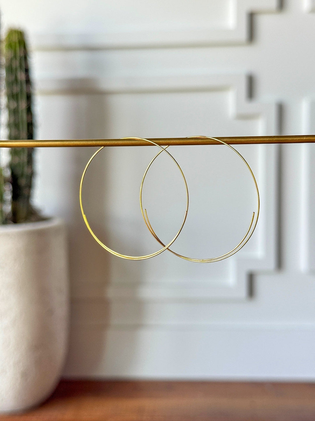 The Gallatin Earrings by Savannah Sevens western life{&}style are large, lightweight gold hoops displayed on a brass rod, with a white wall and potted cactus softly blurred in the background.