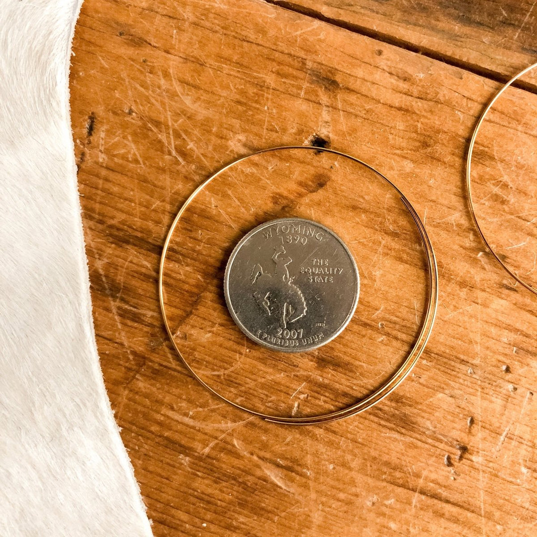 A U.S. quarter rests on wood beside a thin, circular 14K gold plated wire, showing the delicate size of Gallatin Earrings by Savannah Sevens western life{&}style—perfect for lightweight or gold hoop earring styles.