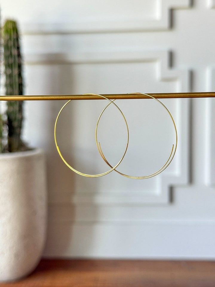 The Gallatin Earrings by Savannah Sevens western life{&}style are large, lightweight gold hoops displayed on a brass rod, with a white wall and potted cactus softly blurred in the background.
