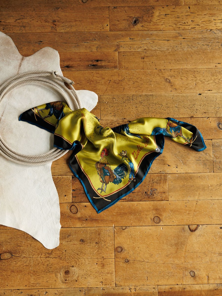 The Gallo Del Cielo Wild Rag from Savannah Sevens western life{&}style, featuring rooster illustrations, rests on a wooden floor next to a coiled rope and a white cowhide rug.