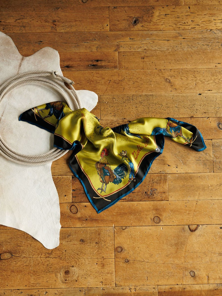 The Gallo Del Cielo Wild Rag from Savannah Sevens western life{&}style, featuring rooster illustrations, rests on a wooden floor next to a coiled rope and a white cowhide rug.