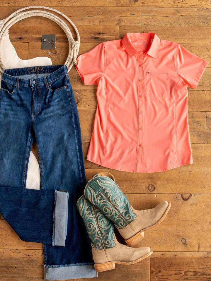 A flat lay of blue jeans, a Galva Ariat Coral Performance Shirt from Savannah Sevens western life{&}style, turquoise and tan cowboy boots, and a white belt on a wooden floor.