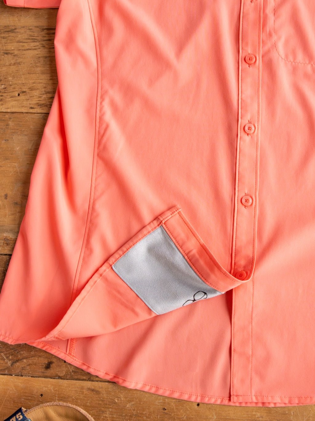 A Galva Ariat Coral Performance Shirt by Savannah Sevens western life{&}style is displayed on a wooden surface, with the lower front hem lifted to show a hidden cleaning cloth sewn inside for convenience.