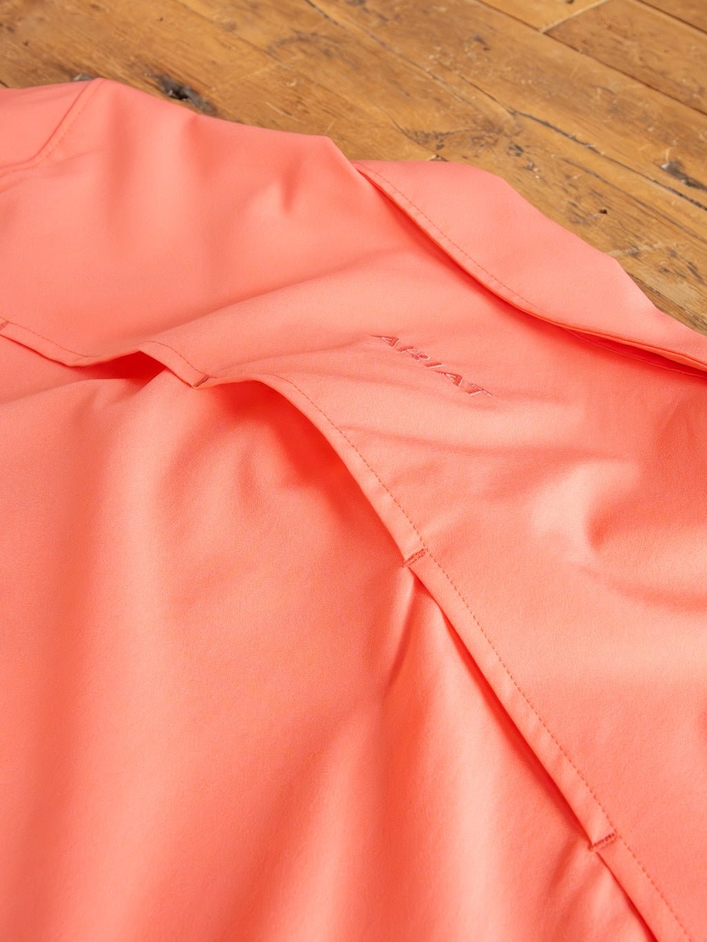 A close-up of the Galva Ariat Coral Performance Shirt by Savannah Sevens western life{&}style, featuring moisture-wicking fabric, vented back panel, subtle embroidered logo, and visible stitching, laid on a wooden surface.