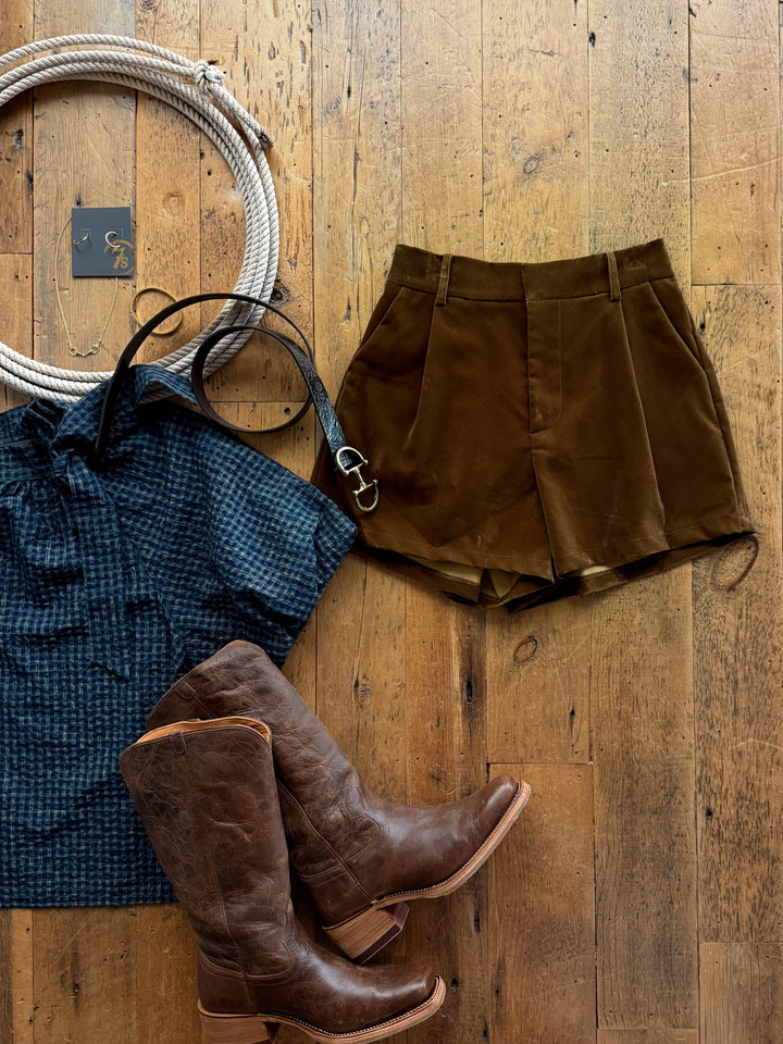 Flat lay of Galva Deep Olive Velvet Shorts, a navy blue plaid blouse, brown leather western boots, a thin belt, a coil of rope, and gold jewelry on a wooden floor.