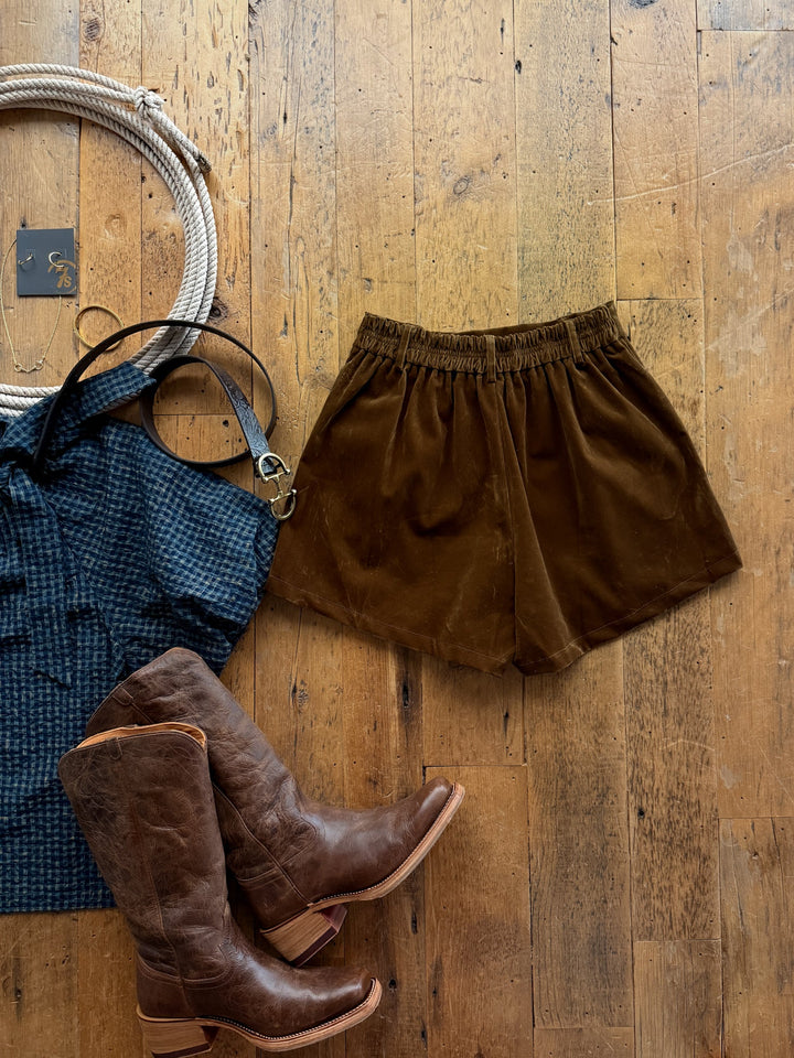 Galva Deep Olive Velvet Shorts laid flat on a wooden floor next to brown leather cowboy boots, a blue patterned top, and jewelry.