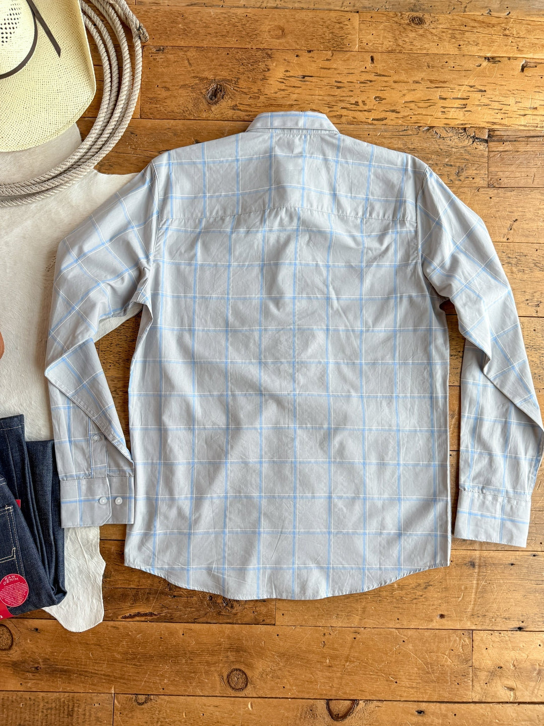 The Garrison {Men's} plaid button-up by Savannah Sevens western life{&}style in light blue, 100% cotton, is laid flat back-up on a wooden floor with a cowboy hat, coiled rope, and dark jeans beside it.