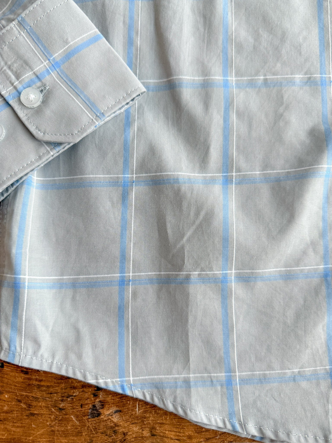 A close-up of The Garrison men's plaid button-up shirt by Savannah Sevens western life&style in light gray with blue and white stripes; 100% cotton for comfort and durability, cuff and sleeve shown on a wooden surface.