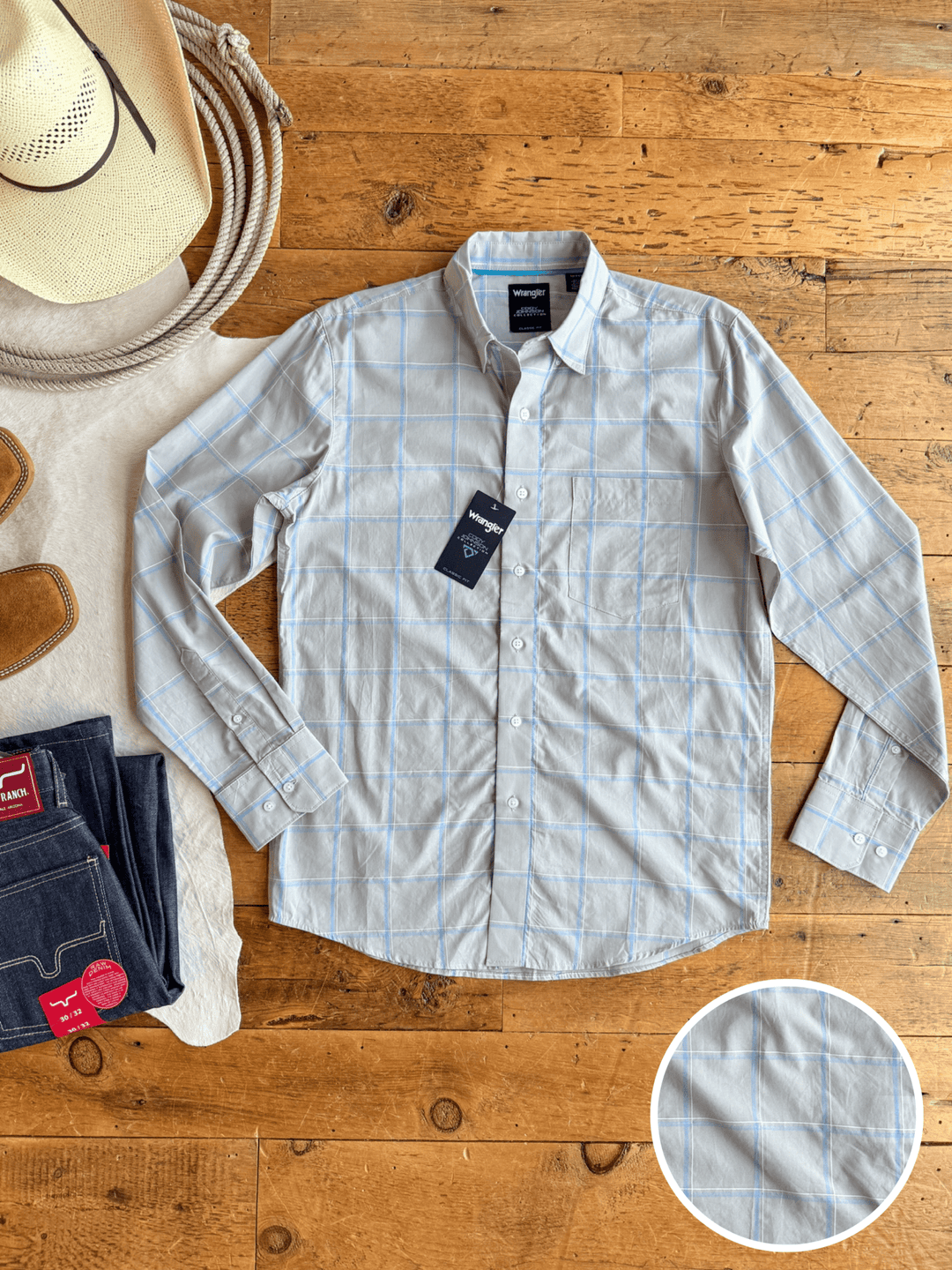 The Garrison men's plaid button-up by Savannah Sevens western life{&}style is laid flat on wood with a cowboy hat, Wrangler boots, and jeans. An inset zooms in on the 100% cotton fabric’s light gray and blue pattern.