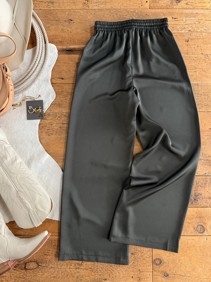The Geneva Olive Satin Wide Leg Pants by Savannah Sevens western life{&}style, featuring a high waist and elastic waistband, are laid out on a wooden floor beside a white cowhide rug, white boots, a beige bag, and a black and gold tag.