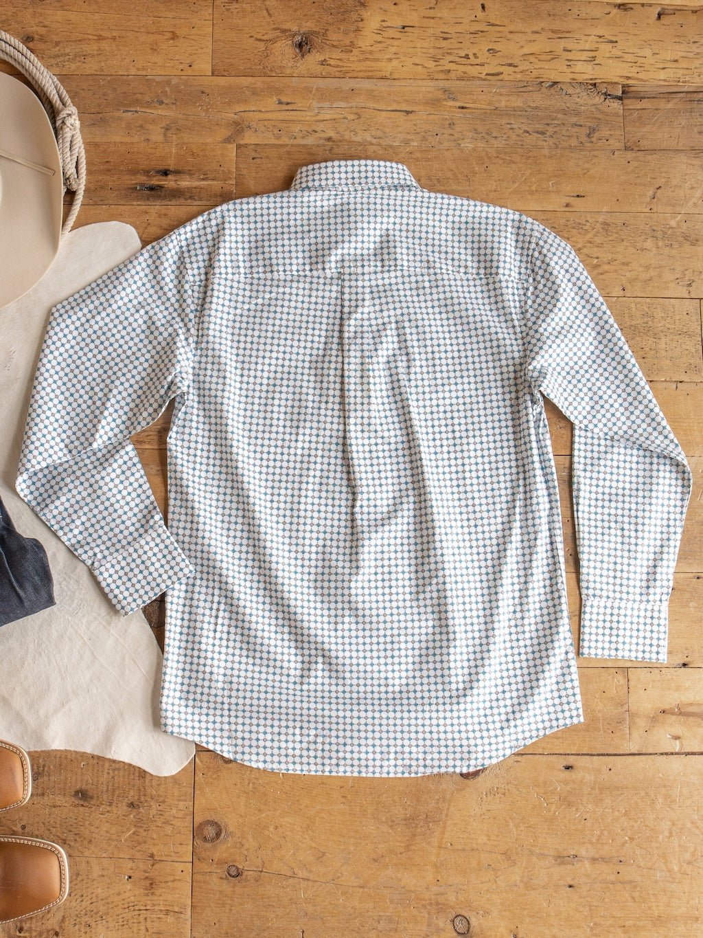 The Georgetown Wrangler Men's Southwest Button Up from Savannah Sevens western life{&}style features a small blue and tan check pattern. It is laid flat, back facing up, on a wooden floor beside a hat and tan fabric.