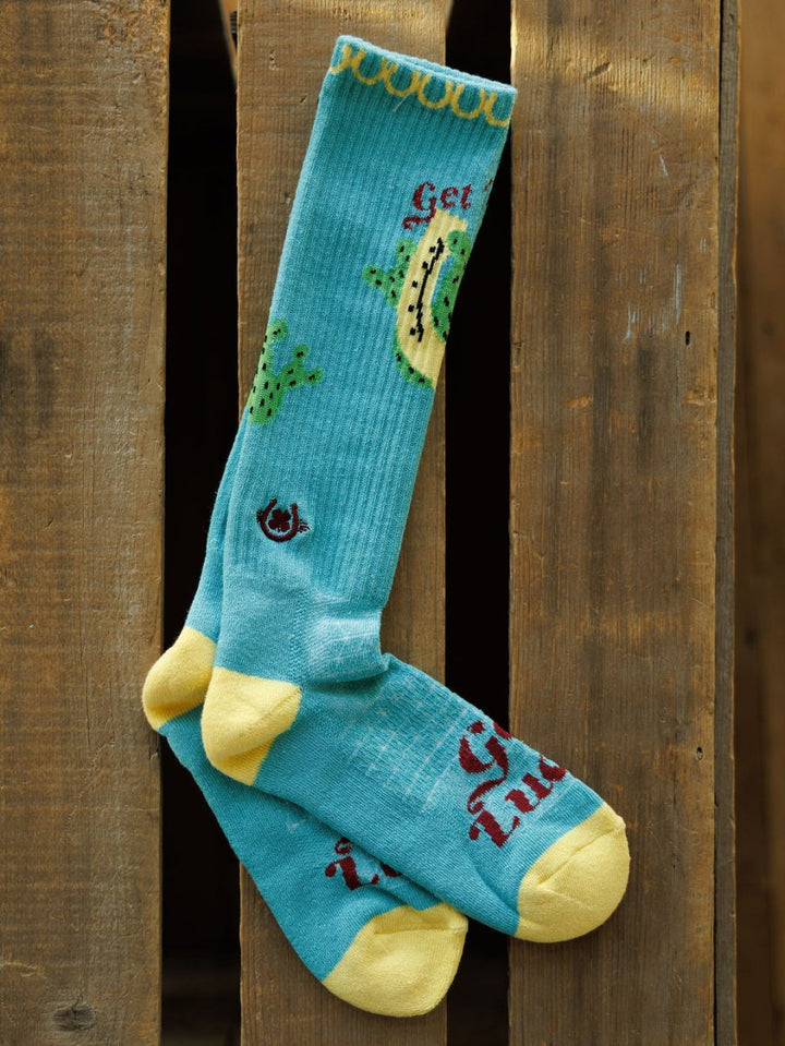 A pair of Get Lucky Crew Socks {Aqua} by Savannah Sevens western life{&}style, blue with yellow toes, heels, and cuffs, avocado and cactus designs, and "Get Tuda" in red text, hanging on a wooden surface.