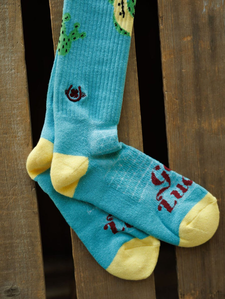 Get Lucky Crew Socks {Aqua} by Savannah Sevens western life{&}style—blue breathable lightweight socks with yellow toes and heels, featuring cactus and horseshoe designs, draped over a wooden surface.