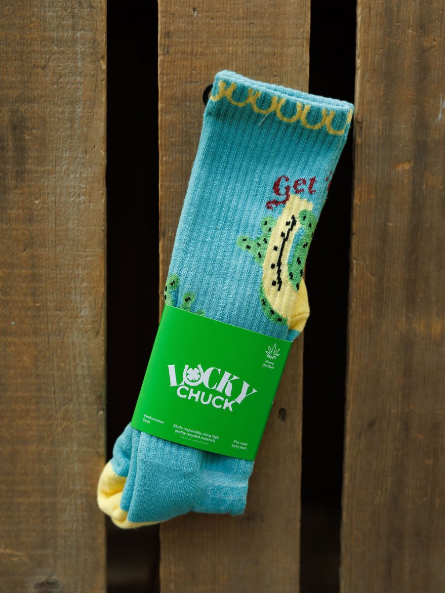 The Get Lucky Crew Socks {Aqua} by Savannah Sevens western life{&}style, featuring yellow accents and a cactus design, are displayed vertically with a green branded label against a rustic wooden background.