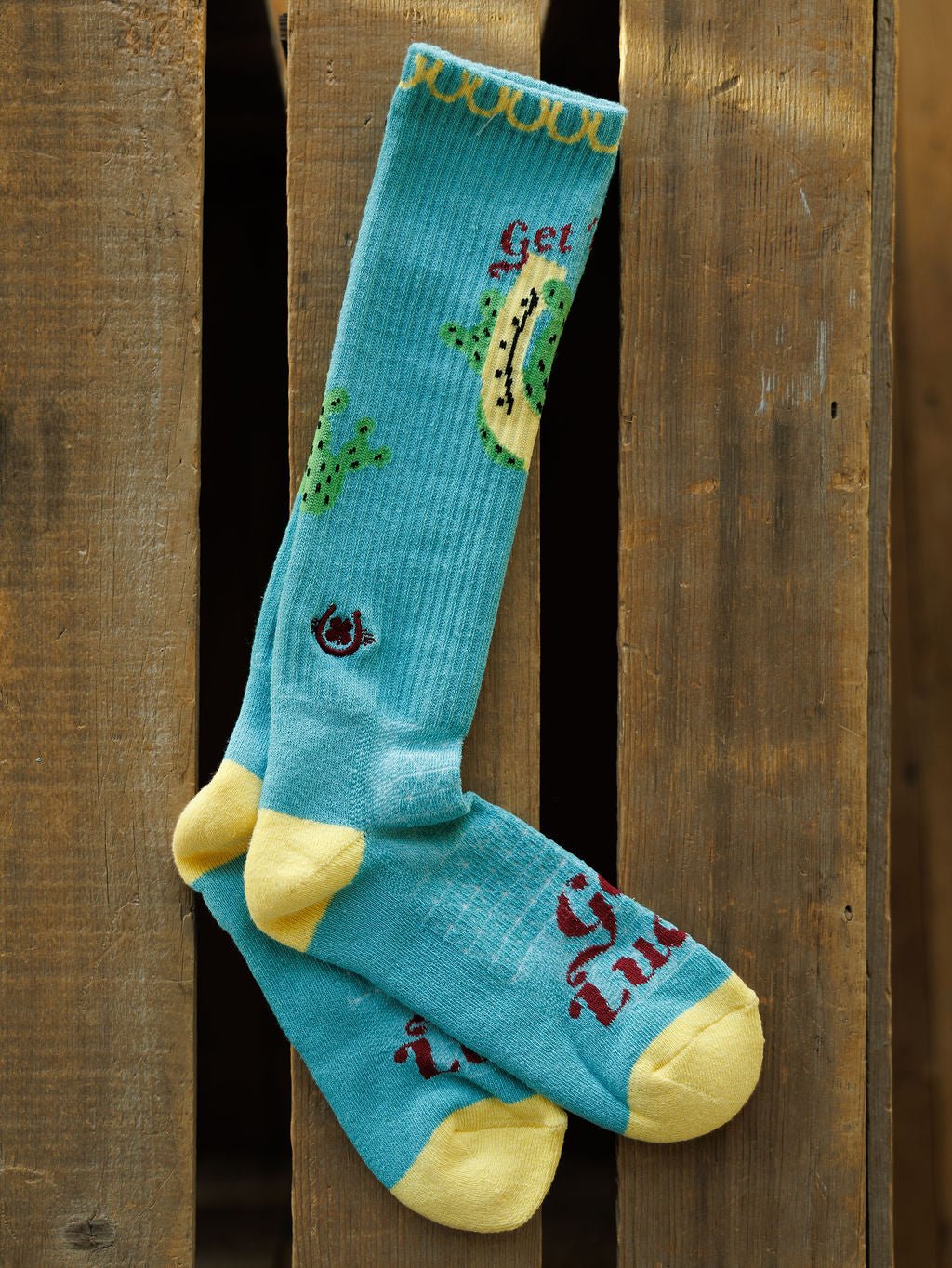 A pair of Get Lucky Crew Socks {Aqua} by Savannah Sevens western life{&}style, blue with yellow toes, heels, and cuffs, avocado and cactus designs, and "Get Tuda" in red text, hanging on a wooden surface.