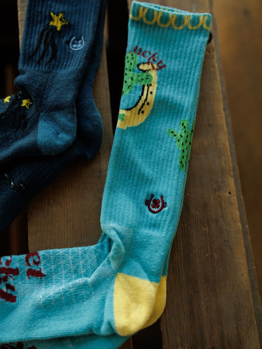 The Get Lucky Crew Socks {Aqua} by Savannah Sevens western life{&}style feature a blue star and moon design and a teal sock with cactus, horseshoe, "lucky" text, plus yellow heel/toe. Breathable and lightweight for comfort.