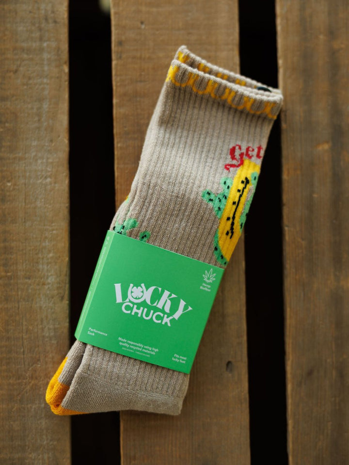 A pair of Get Lucky Crew Socks {Tan} by Savannah Sevens western life{&}style, featuring lightweight breathable fabric with yellow accents and colorful designs, wrapped in a green Lucky Chuck label, displayed on a wooden surface.
