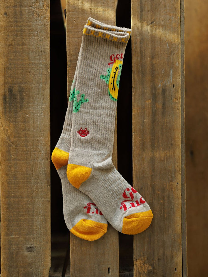 A pair of Get Lucky Crew Socks {Tan} by Savannah Sevens western life{&}style, featuring yellow heels and toes with cactus and chili pepper graphics, hang over a wooden pallet. Red and yellow branding appears near the toes and along the top.