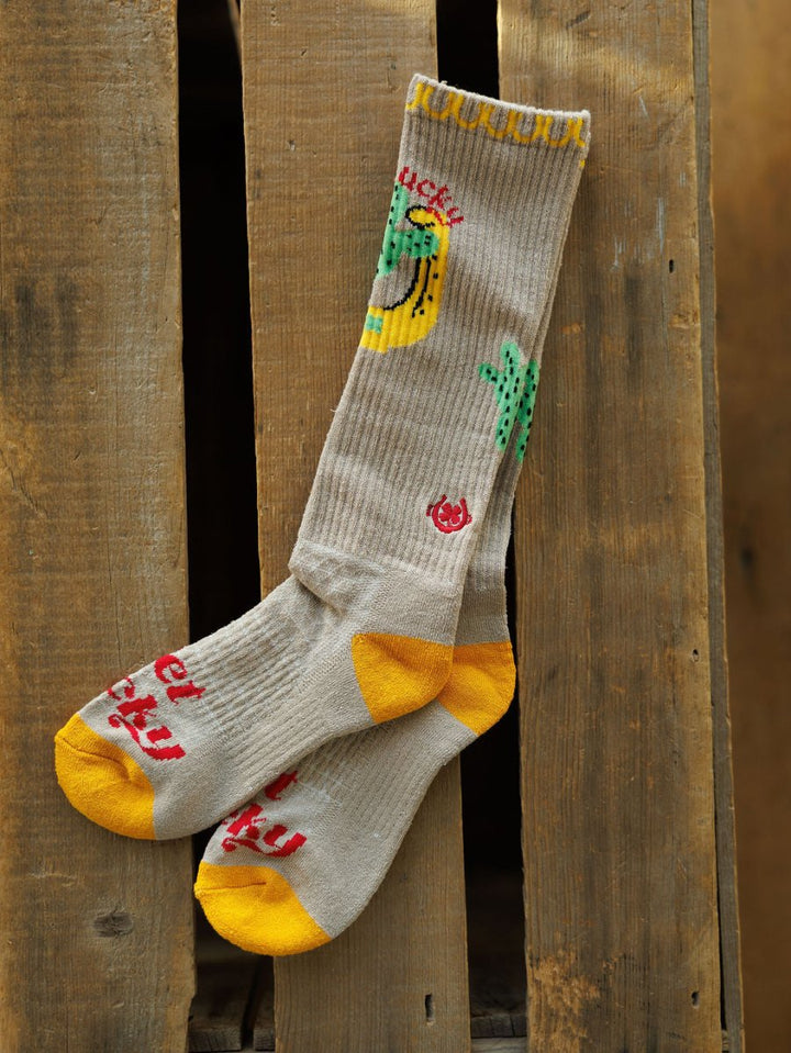 The Get Lucky Crew Socks {Tan} by Savannah Sevens western life{&}style feature beige knee-high design with yellow toes and heels, red sole writing, colorful banana and cactus details, and odor-fighting hemp blend, hanging on wood.