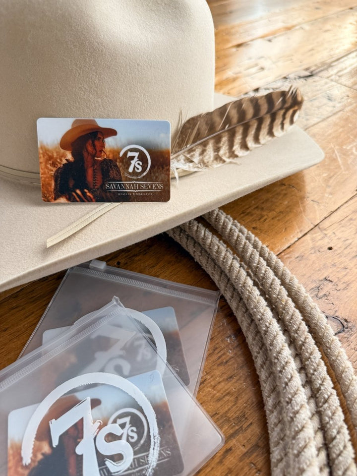 A beige cowboy hat with a feather and a Savannah Sevens western life{&}style Gift Card {Physical Version} on the brim sits by a coiled rope and three more physical gift cards in holders—all displayed on wood, perfect for gifting choice.