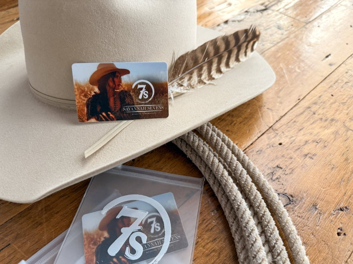 A beige cowboy hat with a feather, a Savannah Sevens western life{&}style Gift Card (Physical Version) featuring a woman in a hat, coiled rope, and Savannah Sevens stickers displayed on a wooden surface.