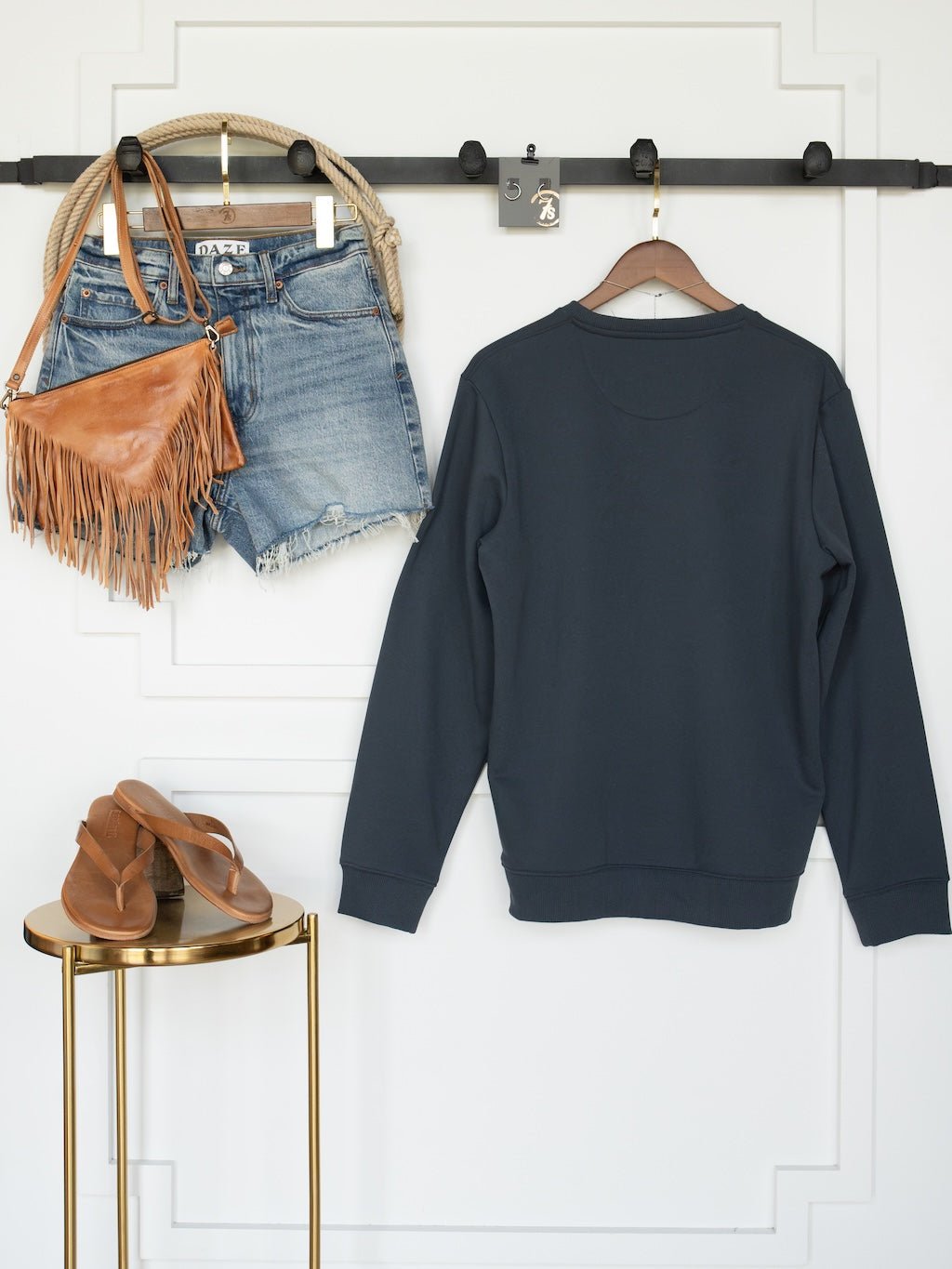 A Gillette Slate Western Graphic Pullover by Savannah Sevens western life{&}style hangs on a hanger, paired with denim shorts and a fringed brown bag beside it, while tan sandals are displayed on a small gold table.