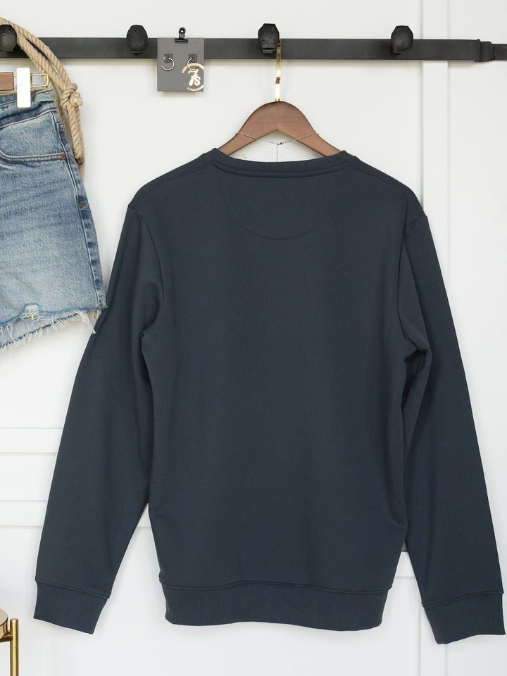 The Gillette Slate Western Graphic Pullover by Savannah Sevens western life{&}style, a dark gray lightweight fleece sweatshirt, hangs on a wooden hanger against a white wall, with denim shorts visible on the left side of the image.