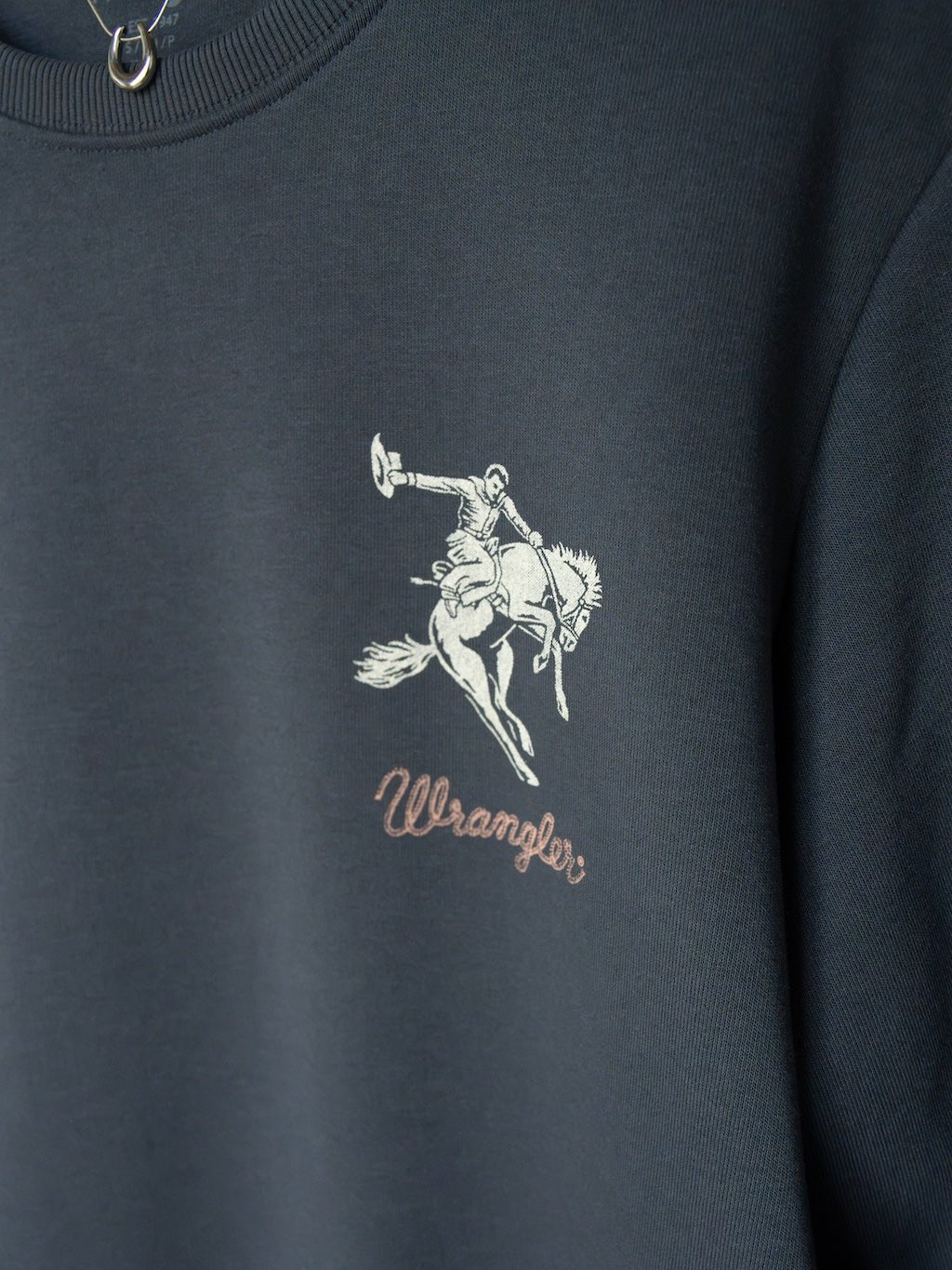Close-up of the Gillette Slate Western Graphic Pullover by Savannah Sevens western life{&}style features a dark western design with a saddle bronc logo depicting a cowboy riding a bucking horse above the word "Wrangler" in script.