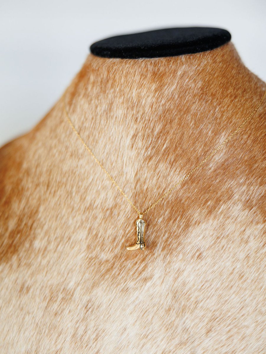 The Savannah Sevens western life{&}style Gold Cowgirl Boot Necklace features a 14K yellow gold plated cowgirl boot, elegantly displayed on a brown and white cowhide bust. Add a touch of western charm to any outfit with this standout jewelry piece.