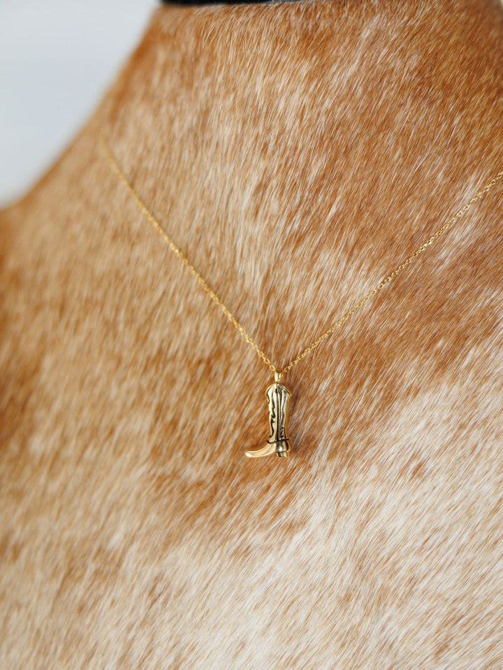 The Savannah Sevens western life{&}style Gold Cowgirl Boot Necklace sits against cowhide bust, showcasing this chic 14K yellow gold plated pendant.