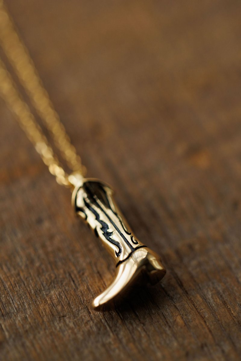 The Gold Cowgirl Boot Necklace by Savannah Sevens western life{&}style, plated in 14K yellow gold with black accents, rests on a wooden surface, capturing the essence of western jewelry.