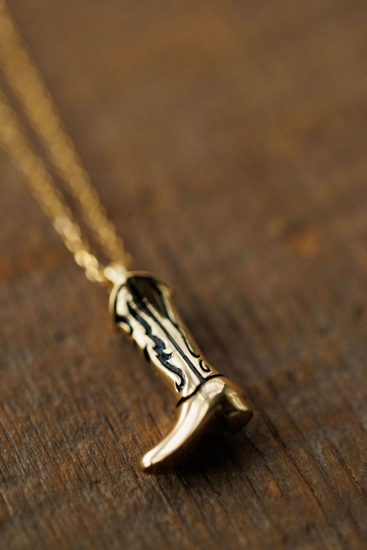 The Gold Cowgirl Boot Necklace by Savannah Sevens western life{&}style, plated in 14K yellow gold with black accents, rests on a wooden surface, capturing the essence of western jewelry.