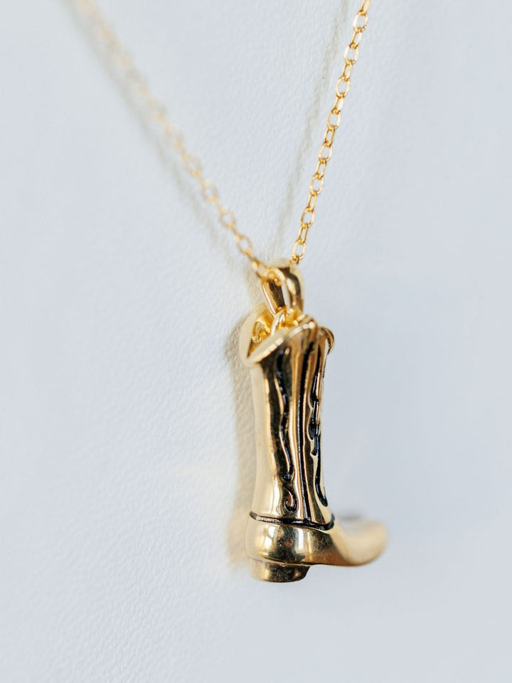 The Gold Cowgirl Boot Necklace by Savannah Sevens western life{&}style features a 14K yellow gold plated cowboy boot design, shown on a white background—a perfect western jewelry accent for your outfit.