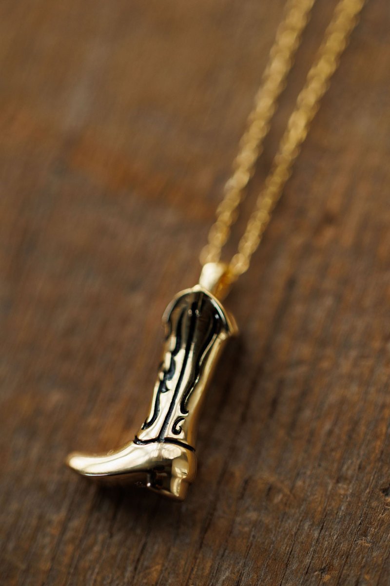 The Gold Cowgirl Boot Necklace by Savannah Sevens western life{&}style showcases a 14K yellow gold plated, engraved boot pendant on a chain, resting on a wooden surface—perfect for adding western flair to any outfit.