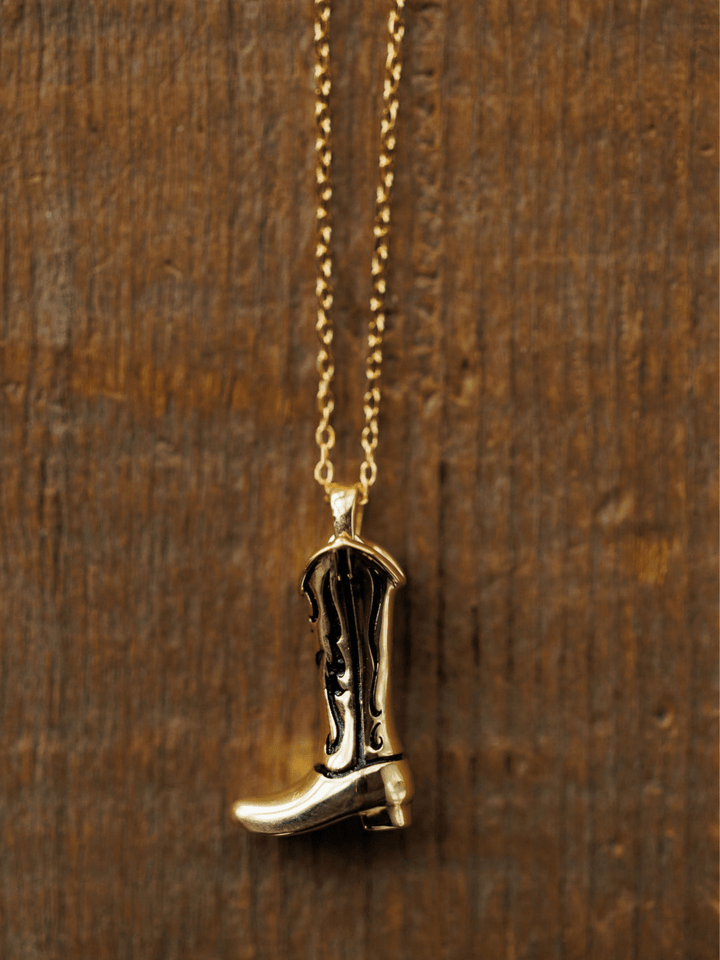 The Gold Cowgirl Boot Necklace by Savannah Sevens western life{&}style features 14K yellow gold plating and black detailing—a perfect piece of western jewelry for cowgirl style lovers. Displayed against a rustic wooden background.