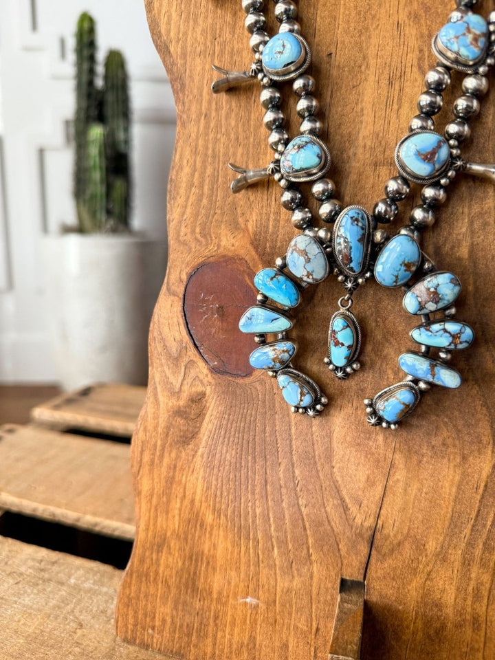 The Golden Hills Necklace & Earrings Set by Savannah Sevens western life&style, featuring turquoise accents and Navajo pearls, hangs on a wooden display stand with a blurred potted cactus in the background.