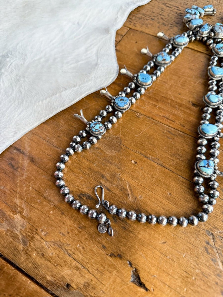 The Golden Hills Necklace & Earrings Set by Savannah Sevens western life&style, featuring turquoise stones and silver beads, is displayed on a wooden surface beside a piece of white fur.