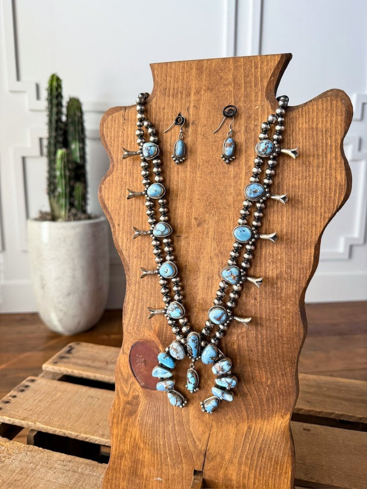 The Golden Hills Necklace & Earrings Set by Savannah Sevens western life{&}style, featuring turquoise and Navajo pearls, is displayed on a wooden bust atop a crate with a tall cactus in a white pot in the background against a white-paneled wall.